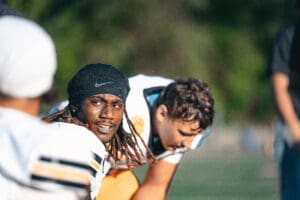 Football players in uniform, one smiling in foreground during practice, green field in background.
