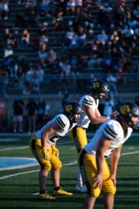 Football players in yellow uniforms ready for play on field during sunny day game, with crowd in background.