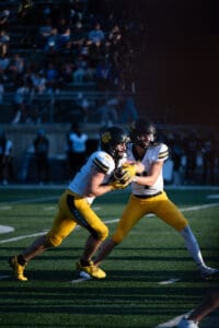 Football players in yellow uniforms executing a handoff play during a game on a sunny day.