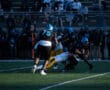 High school football game action under the lights, players tackling on the field, crowd watching in background.