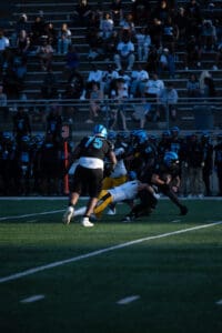 High school football game action under the lights, players tackling on the field, crowd watching in background.