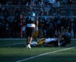 High school football game action with players tackling, fans watching in background.