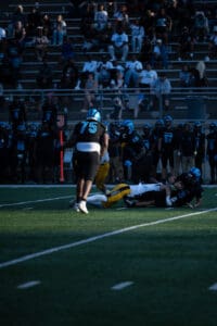 High school football game action with players tackling, fans watching in background.
