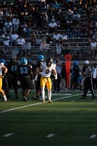 Football game in progress with players in black and yellow uniforms on the field, crowd watching in the stands.