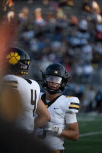 Two football players in helmets and uniforms discussing during a game on a field, with spectators blurred in the background.