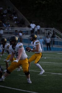 Football quarterback holds ball ready to throw, teammates protect, during a game on a green field.