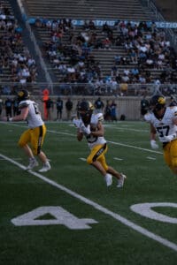 Football players in yellow uniforms running on the field during a game, with stadium crowd in the background.