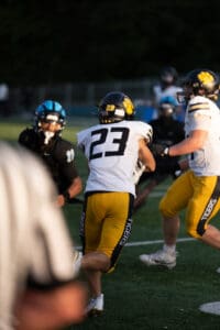 Football game action with player 23 carrying the ball, pursued by opponents on a sunny field.