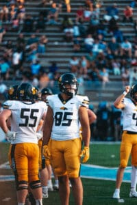 Football players in yellow and white uniforms on the field during a game with a seated crowd in the background.