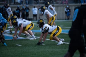 Football players on the line of scrimmage, yellow and black team facing blue and black team. Game underway.