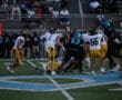 High school football game action with players tackling on the field, fans watching, and bright team uniforms visible.
