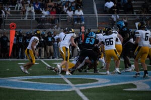 High school football game action with players tackling on the field, fans watching, and bright team uniforms visible.
