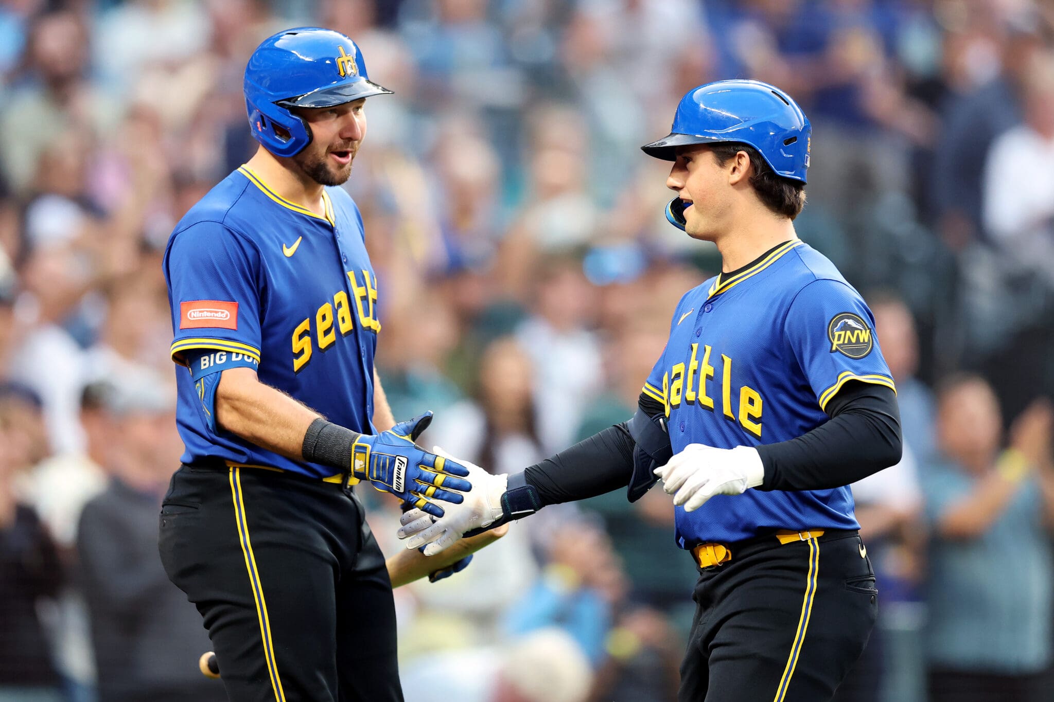 Two Seattle baseball players in blue jerseys celebrating with a handshake on the field.