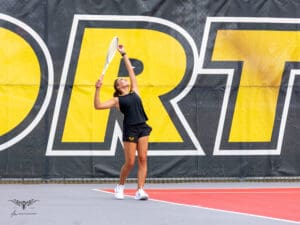 Tennis player serving on court with large yellow lettering background, wearing black and holding a white racquet.