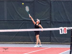 Tennis player in a black outfit serves during a match on a red court with a visible score of 1-0.