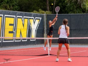 Two women playing a competitive tennis match on a bright red court, one serving the ball.