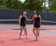 Two female tennis players shake hands on a red court during a doubles match, wearing black outfits and visors.