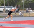 Tennis player in action on court, backhand swing, red and gray court, spectators in background.
