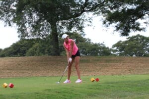 Golfer in pink preparing to swing on a lush course. Trees in background, wearing cap and gloves.
