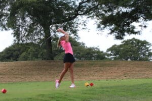 Golfer in pink outfit finishes powerful swing on green course against tree backdrop.