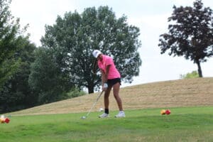 Golfer in pink shirt prepares to swing on a lush golf course, surrounded by trees on a sunny day.