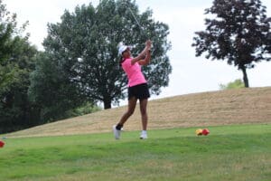 Golfer in pink shirt swinging club on green fairway surrounded by trees on a sunny day.