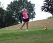 Golfer in pink shirt swings club on lush green fairway, with trees and distant player in background.