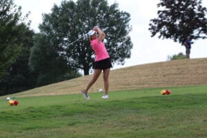 Golfer in pink shirt swings club on lush green fairway, with trees and distant player in background.