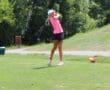 Golfer in pink shirt and cap swings on lush green course with trees in background.