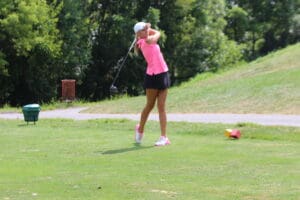 Golfer in pink shirt and cap swings on lush green course with trees in background.