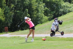 Golfer in pink shirt swinging club on lush course with golf bag cart nearby.