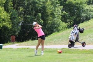 Golfer in pink top swinging club on sunny day, with golf cart on course.