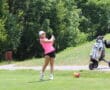 Golfer in pink shirt swings club on a sunny course, with a golf cart and lush green trees in the background.