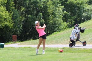Golfer in pink shirt swings club on a sunny course, with a golf cart and lush green trees in the background.