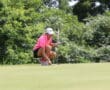 Golfer lines up putt on the green, wearing pink shirt and white cap, surrounded by lush trees.