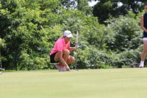 Golfer lines up putt on the green, wearing pink shirt and white cap, surrounded by lush trees.