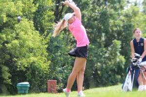 Golfer in pink shirt swings club on a sunny course surrounded by trees.