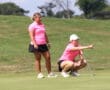 Two women in pink shirts strategize on a sunny golf course, one squatting and pointing, focused on the game.
