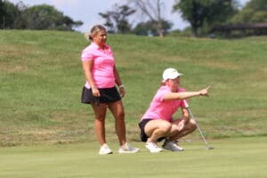 Two women in pink shirts strategize on a sunny golf course, one squatting and pointing, focused on the game.