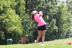 Golfer in pink shirt swings club on lush green course, surrounded by trees and bright sunlight.
