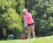 Golfer in pink shirt preparing to swing on a sunny day, surrounded by green trees.