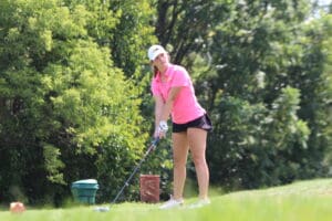 Golfer in pink shirt preparing to swing on a sunny day, surrounded by green trees.