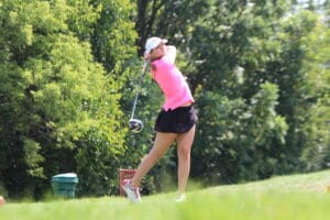 Golfer in pink shirt and black skirt swinging on a lush, green course.