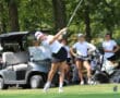 Golfer swings club on green near golf cart, watched by teammates on a sunny day.