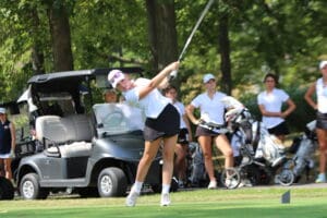 Golfer swings club on green near golf cart, watched by teammates on a sunny day.