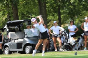 Golfer tees off at lush golf course, joined by teammates and golf cart under trees.