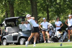 Golfer swings on sunny course with golf cart and teammates in background.