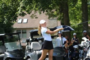 Golfer swings club near parked golf carts, sunlight filtering through trees in the background.