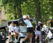 Golfer swings club on a sunny day, with teammates and golf carts in the background on a lush green course.