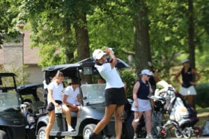 Golfer swings club on a sunny day, with teammates and golf carts in the background on a lush green course.